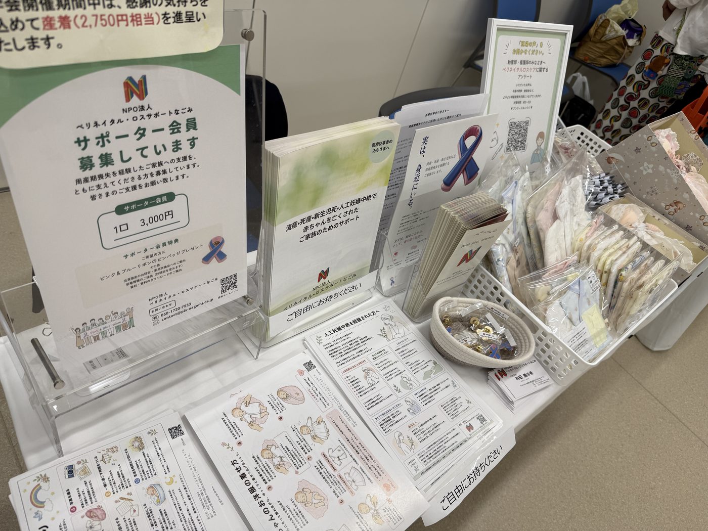 ブースの展示物を近くから写した様子。サポーター会員募集の案内板、ご家族向けのパンフレット、ピンク＆ブルーリボンのピンバッジや産着などが並んでいる。