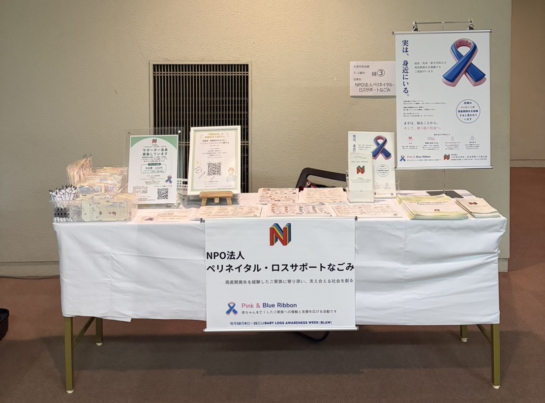 日本母性衛生学会に出展したNPO法人ペリネイタル・ロスサポートなごみのブースの様子。長机にパンフレットや資料が並び、ピンク＆ブルーリボンの啓発ポスターが掲示されている。